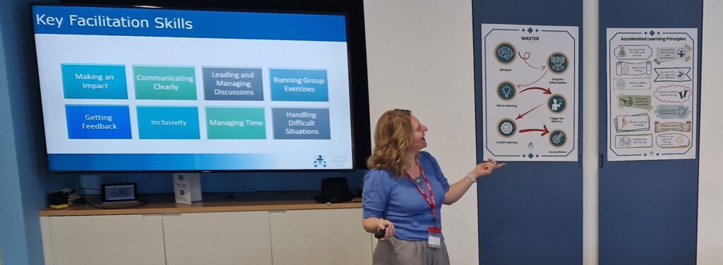 Sheridan Webb presenting to a group with slides explaining key facitation skills and posters showing adult learning principles