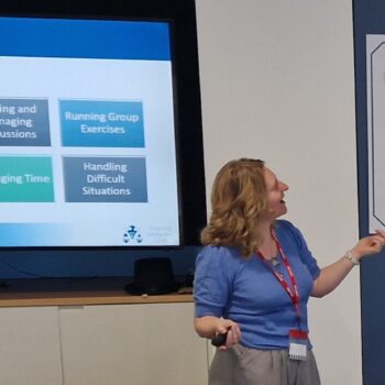 Sheridan Webb presenting to a group with slides explaining key facitation skills and posters showing adult learning principles